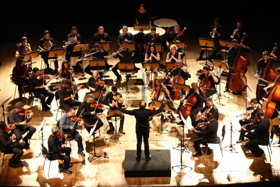 Músicos da orquestra são regidos pelo maestro Luiz Petri, de costas e ao centro da imagem. #pratodosverem. 