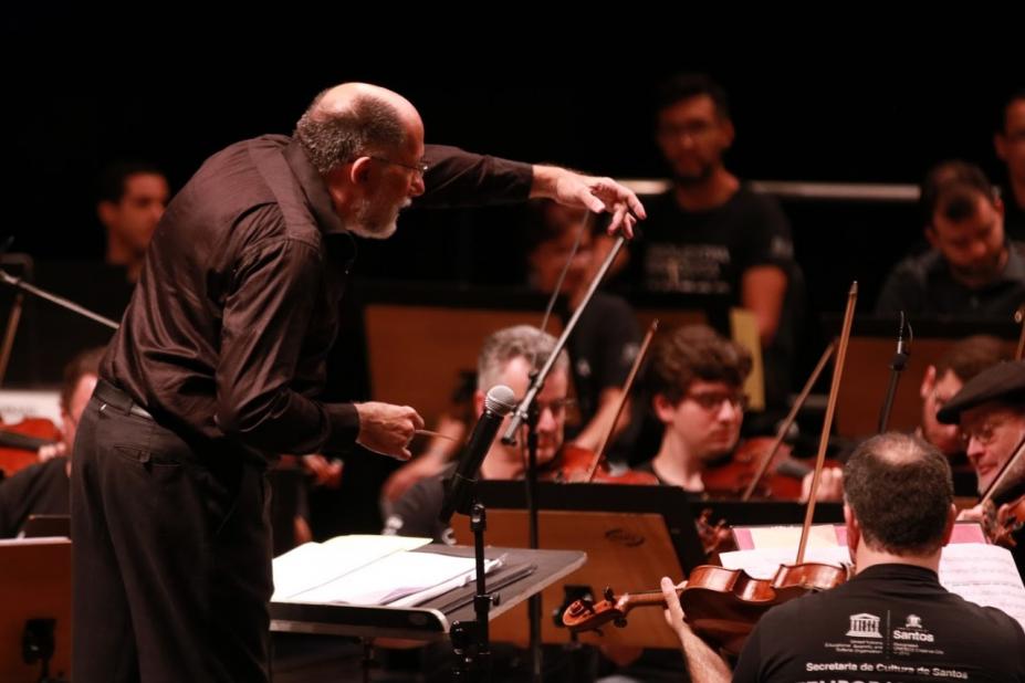 maestro em primeiro plano está regendo orquestra ao fundo. #Paratodosverem