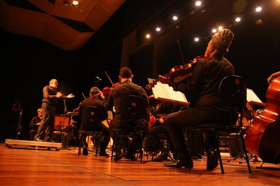 músicos sentados tocando com maestro ao fundo num teatro. #paratodosverem 