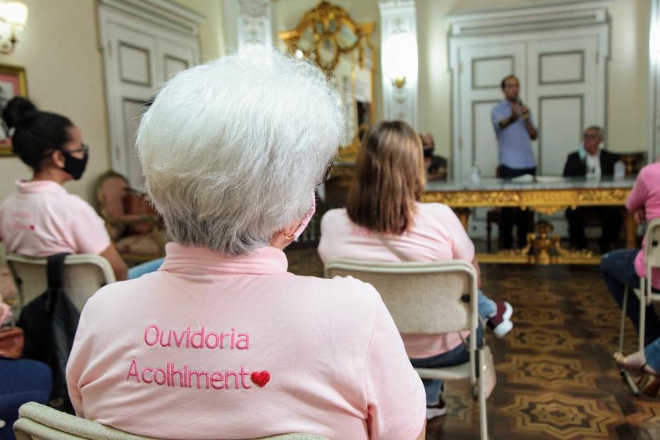 Em primeiro plano, mulher está sentada, de costas. Ela veste uma blusa onde se lê OUvidoria Acolhimento. Ao fundo, a mesa de autoridades. #Paratodosverem