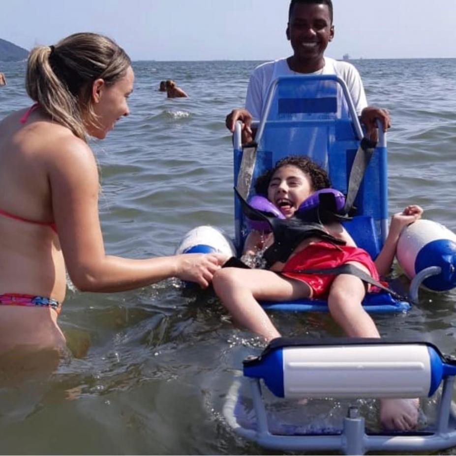 menina está em cadeira anfíbia conduzida por um jovem. Ao lado da menina, uma mulher a acompanha. Todos sorriem. #Paratodosverem