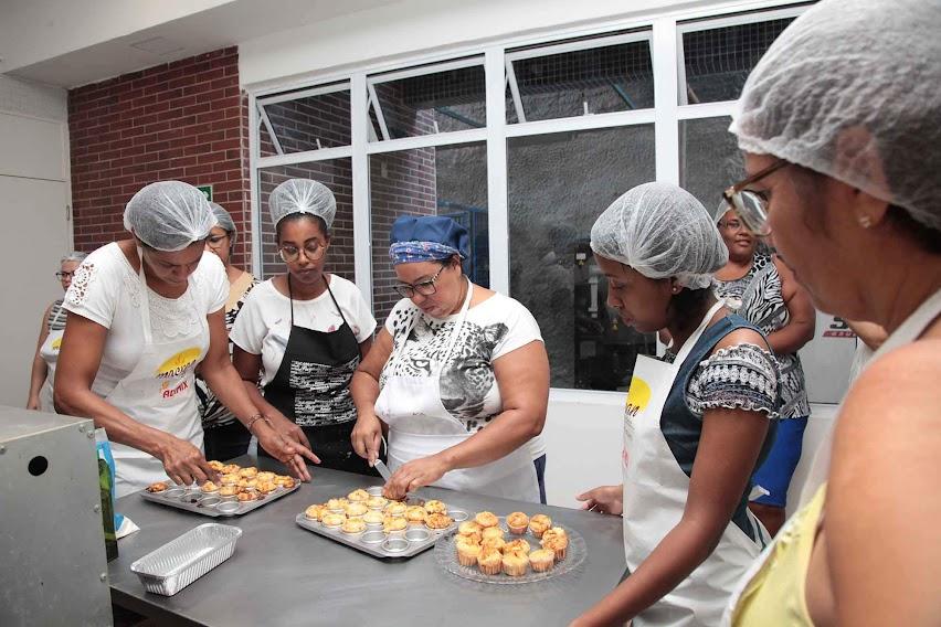 mulheres estão reunidas em torno de uma mesa desenformando empadinhas. #paratodosverem 