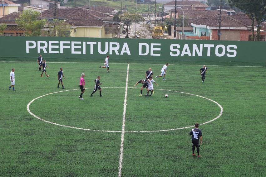 jogo de veteranos no campo renovado. Ao centro, há uma disputa de bola. Ao fundo, se lê: Prefeitura de Santos. #paratodosverem