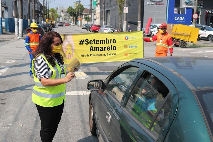 Mulher entrega panfleto a motorista e ao fundo faixa do setembro amarelo #paratodosverem