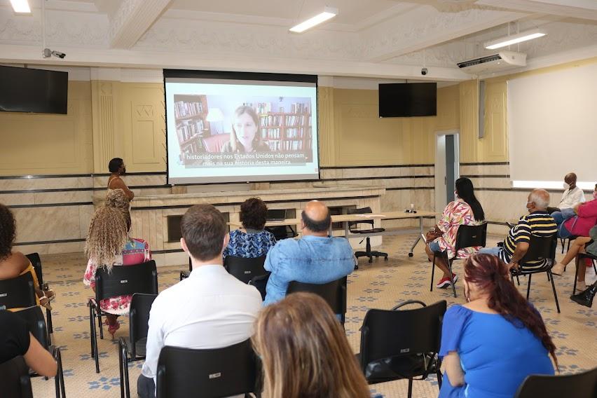 pessoas estão sentadas em salão, de costas para foto, e assistindo projeção em telão ao fundo. Uma mulher fala a todos ao lado da projeção. #paratodosverem