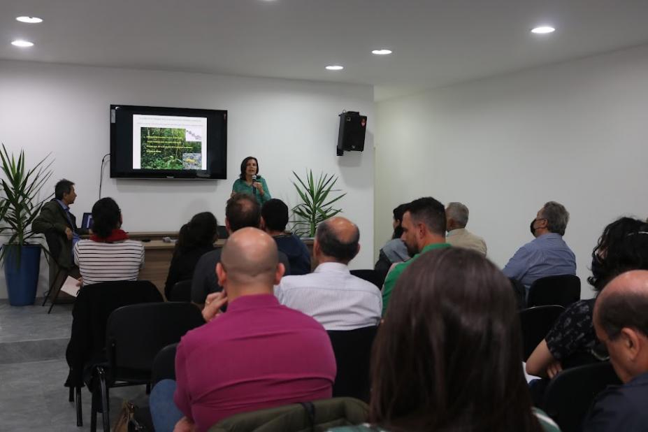 pessoas estão sentadas em auditório de costas para foto. Ao fundo, uma mulher fala ao público. Atrás dela há um monitor com imagem projetada. #paratodosverem