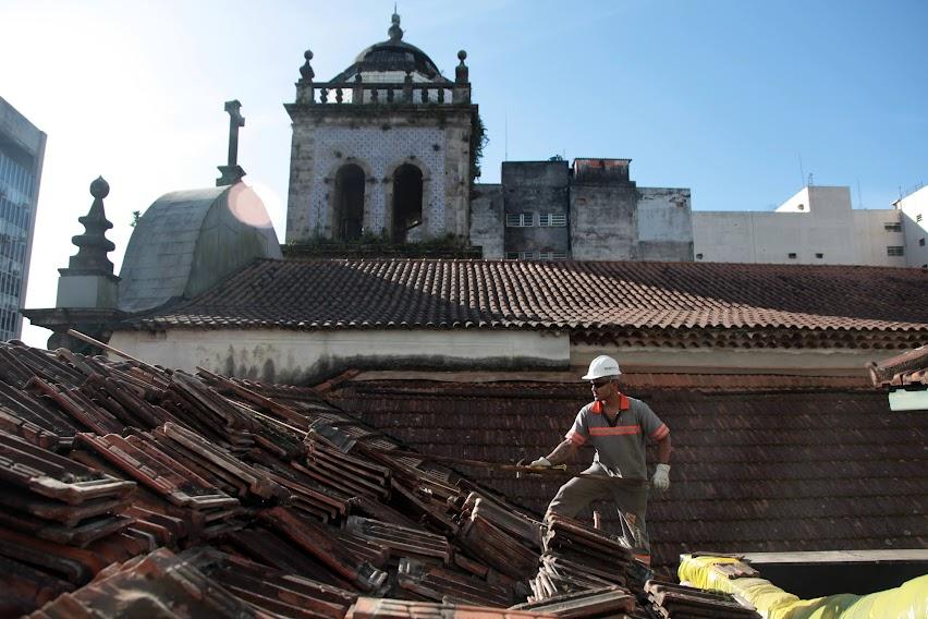 operário está em sobre o telhado de um prédio retirando as telhas. Ao fundo se vê as torres de um conjunto de igrejas. #paratodosverem