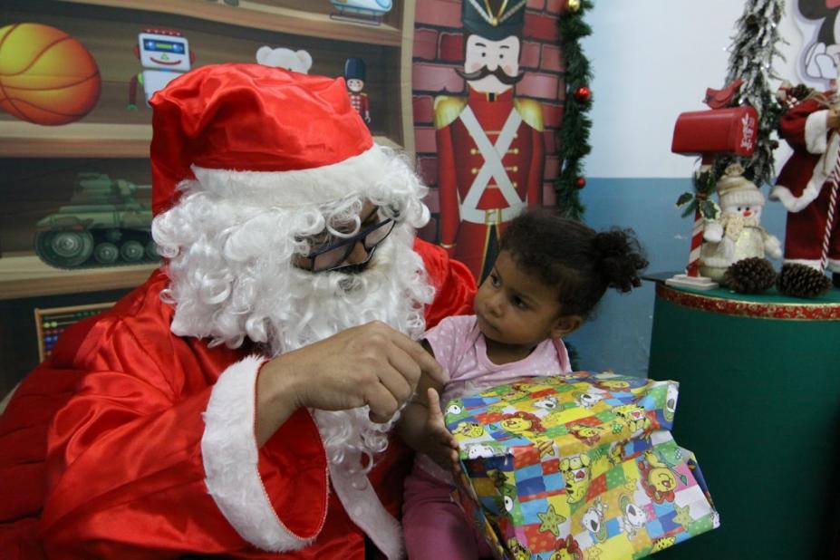 Criança olha curiosa para o papai noel, sentada no colo dele e segurando um presente. #paratodosverem