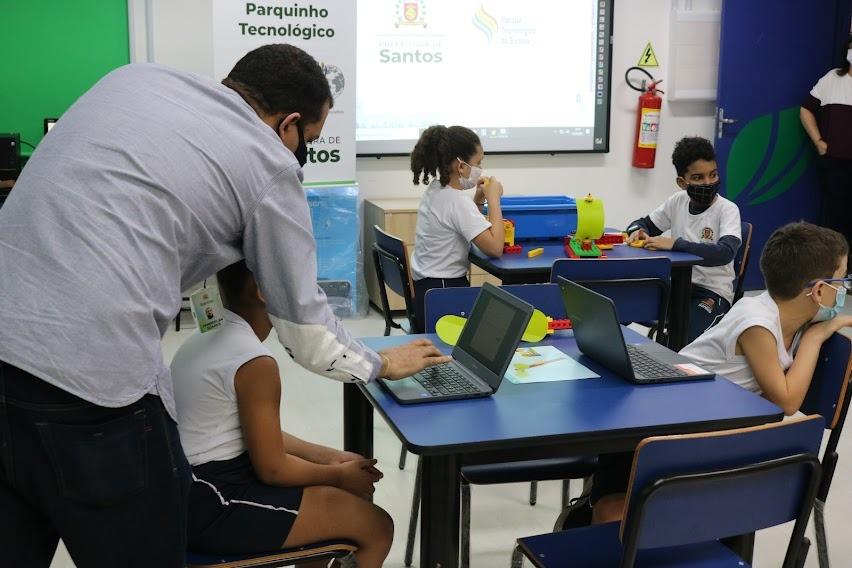 crianças estão sentadas em sala, diante de notebooks. Em primeiro plano, à esquerda, um homem, de costas, tecla no notebook ao lado de uma criança. #paratodosverem