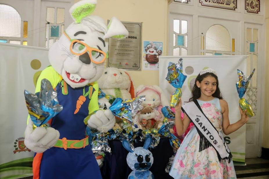 Pessoa vestida com fantasia de coelho e a miss santos mirim estão à frente de mesas com bonecos e ovos de chocolate. Eles dois também seguram ovos nas quatro mãos. #paratodosverem