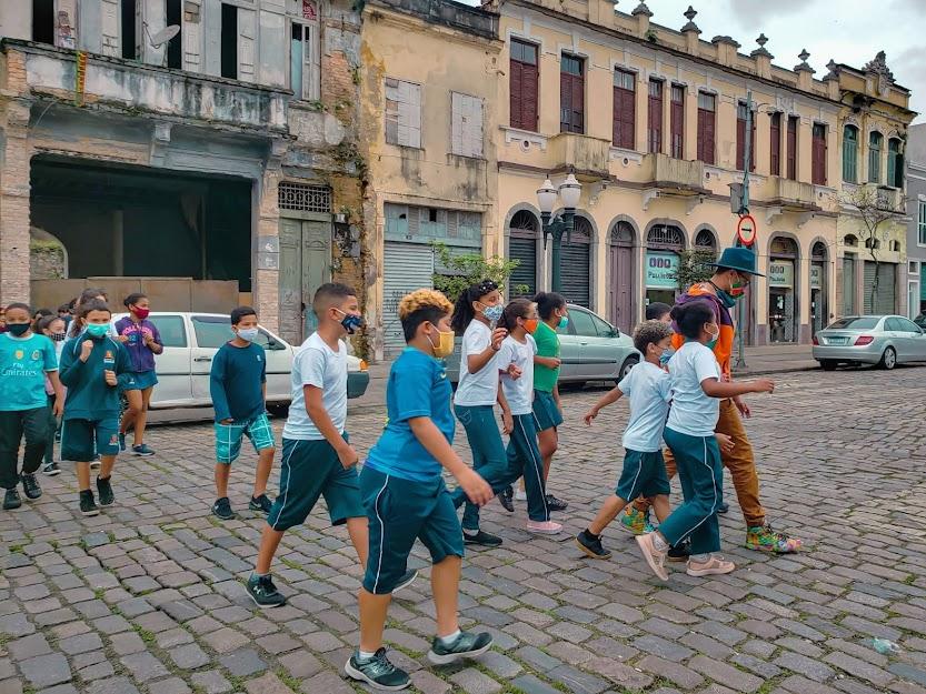 Crianças caminham por rua de paralelepípedo e casas antigas ao fundo. #paratodosverem