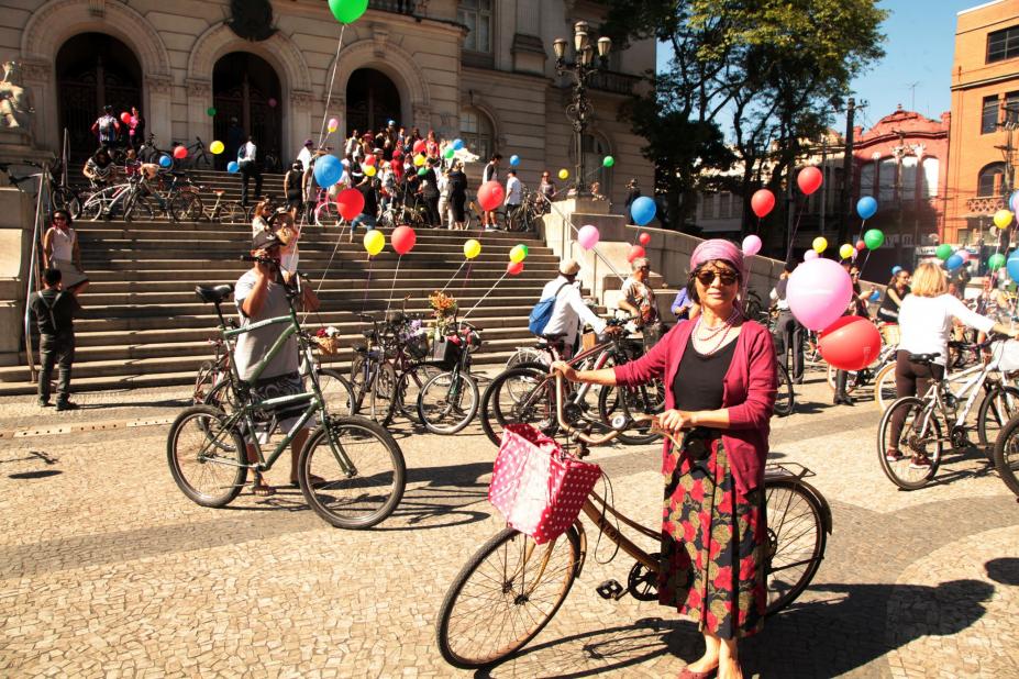 Ciclistas ocupam praça e a escadaria da Praça Mauá. A maioria das bicicletas está decorada com balões coloridos. Em primeiro plano há uma mulher vestida à moda antiga. Ela está em pé à frente de sua bike. #Pracegover