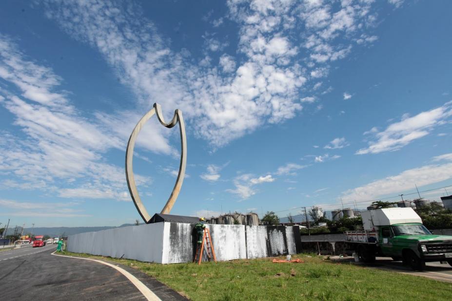 Escultura do Peixe ao fundo, em canteiro central de via. Peça está cercada, na base, por tapume. #Paratodosverem