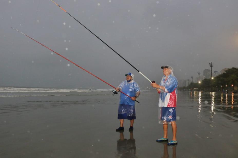 dois homens estão com capa de chuva, em dia chuvoso, pescando na beira do mar. Eles seguram as varas. #paratodosverem