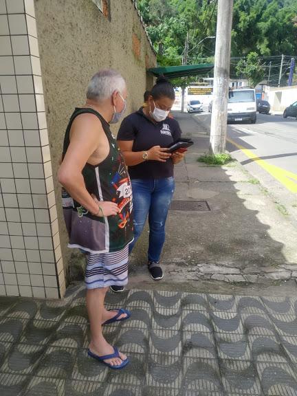 Mulher usando camiseta com identificação da Prefeitura está segurando bloco de notas e conversa com um homem na rua. #Paratodosverem