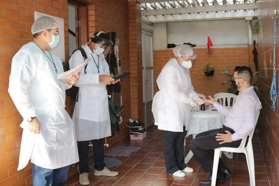 Homem está sentado fazendo teste rápido. Três pesquisadores estão em pé. #Paratodosverem