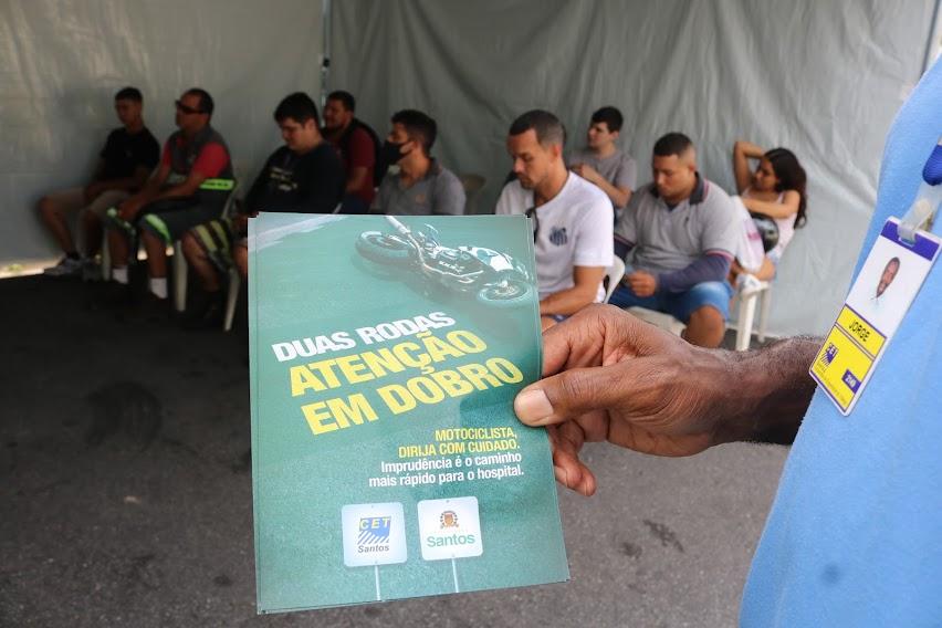 mão segura folheto onde se lê: duas rodas atenção em dobro. Ilustração é de uma moto no chão. ao fundo da foto estão pessoas sentadas sob uma tenda. #paratodosverem