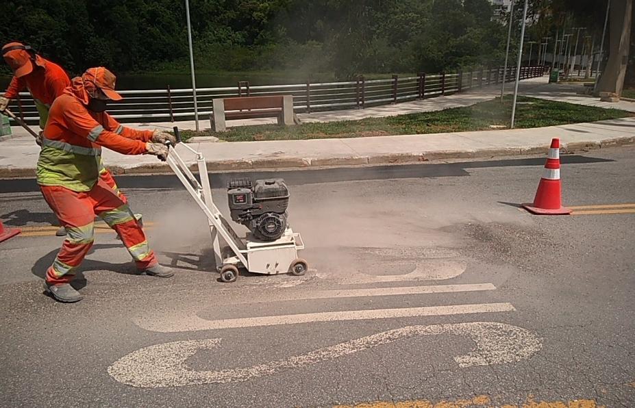 Homem utiliza máquina para apagar pintura antiga em pista. A via está sinalizada por um cone. #Paratodosverem