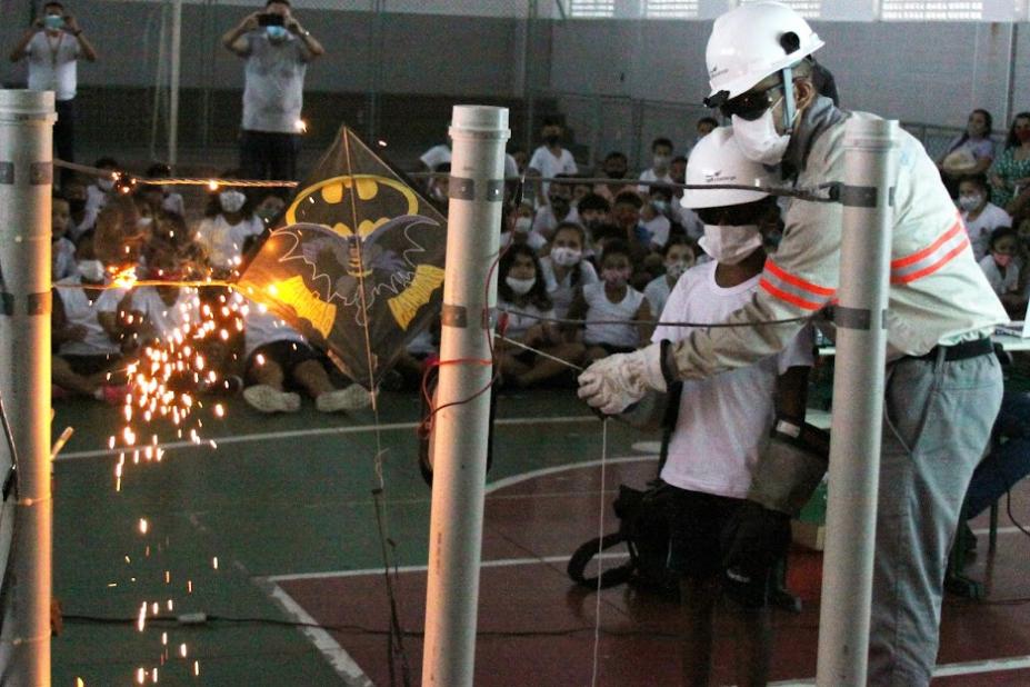 orientador da cpfl uniformizado esá ao lado de menino, ambos protegidos com luvas, capacete e outros itens. Juntos eles puxam a rabiola da pipa presa a um fio e aparece uma faísca. #paratodosverem