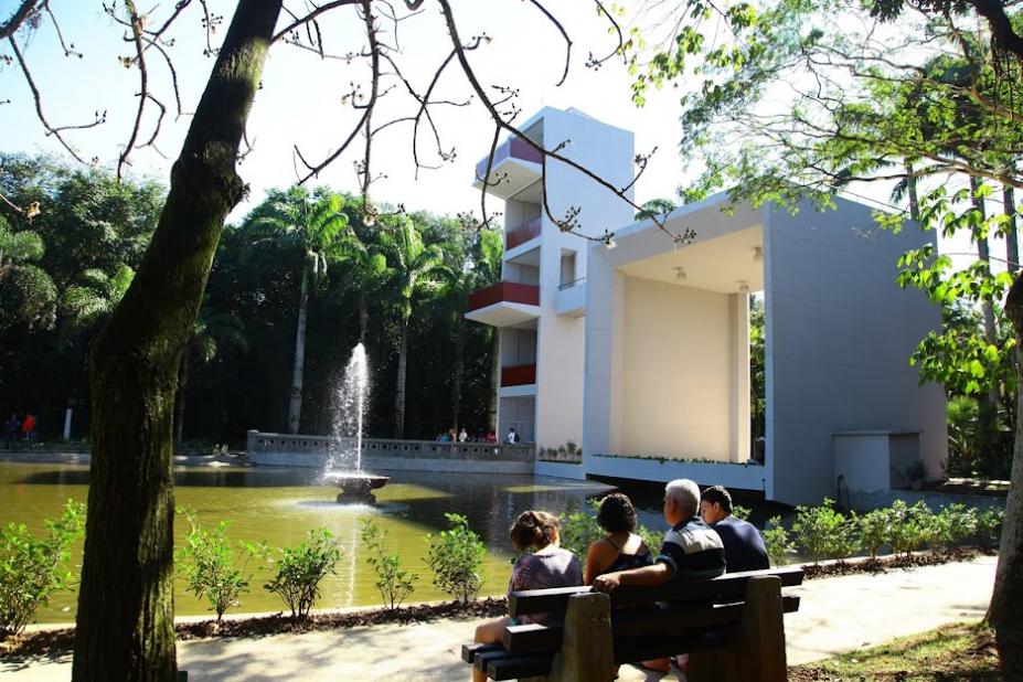 área verdade, com árvore e primeiro plano, banco com quatro pessoas ao lado. Elas estão olhando para um lago com um jato d'água ao centro, que tem, à direita, um mirante de três pavimentos. #paratodosverem