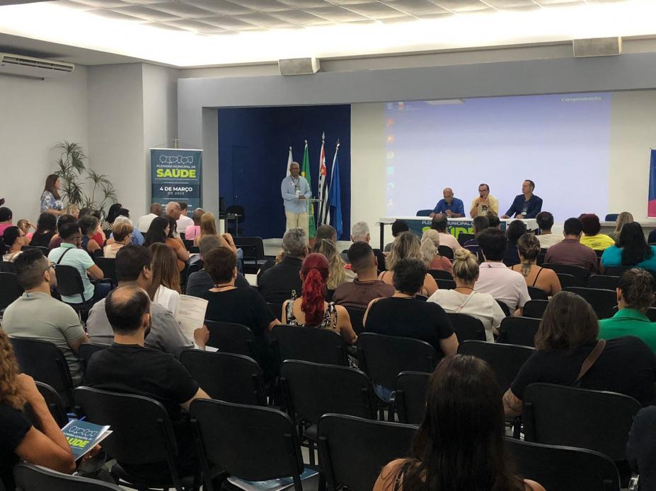 auditório com pessoas sentadas e umas na mesa, falando ao público. #paratodosverem