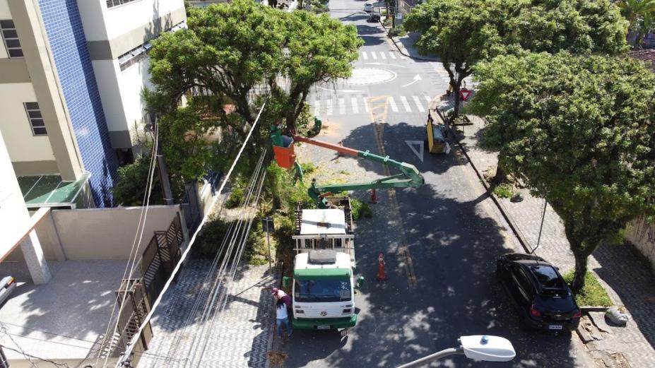 caminhão próprio para poda, com braço articulado e cesto. Homem está dentro do cesto com serra para poda de árvore. Foto feita por drone, com vista do alto para a copa das árvores