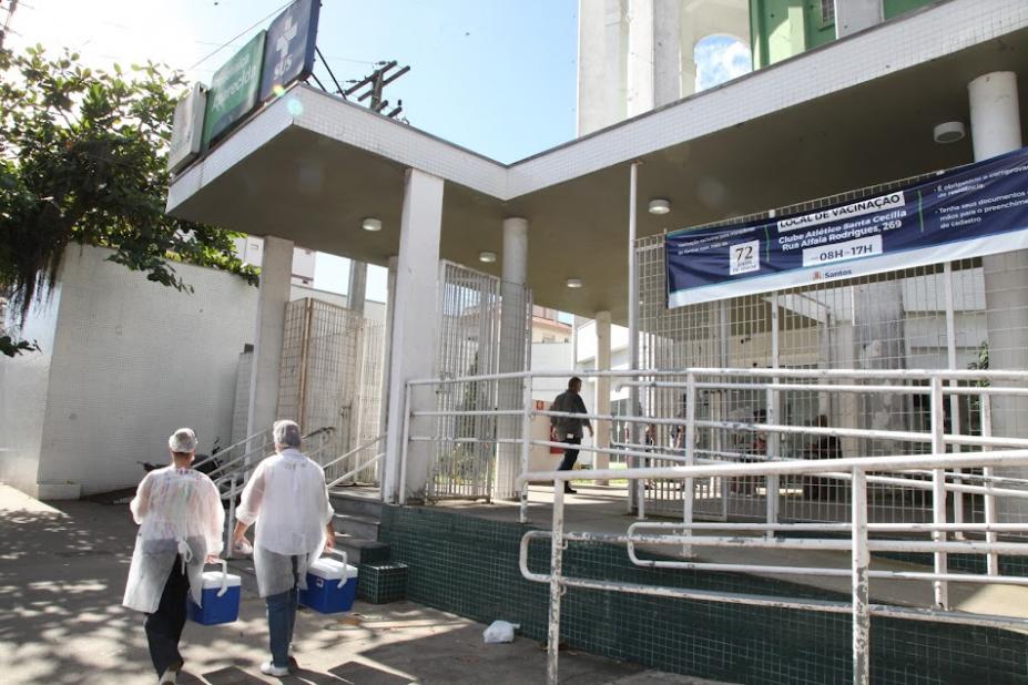 fachada de policlínica com duas pessoas usando avental e segurando coolers entrando. #paratodosverem