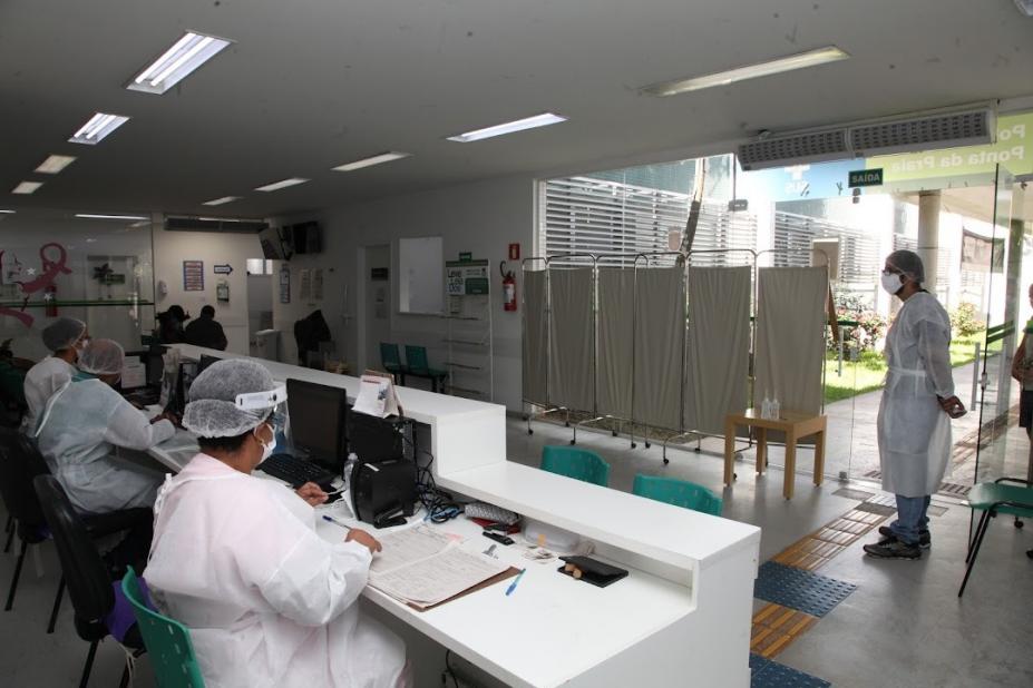 recepção de policlínica com pessoas sentadas atrás de balcão. Elas usam avental, touca e máscara. Ambiente tem outras pessoas em pé. #paratodosverem