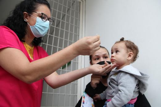 Criança, no colo da mãe, recebe dose de vacina por profissional de saúde. #pratodosverem