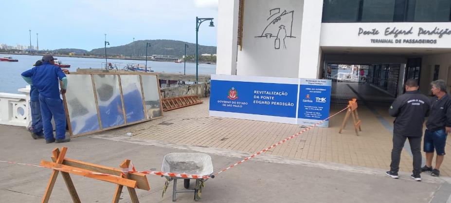 fachada da ponte com placa na frente, cavalete e carrinho de mão na calçada. Homens segurando placa. #paratodosverem