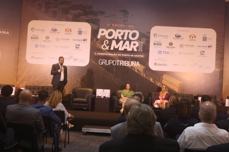 prefeito fala em palco. Ele está ao lado esquerdo da foto em pé. Do lado esquerdo, duas mulheres sentadas. A plateia assiste. Ao fundo, um painel onde se lê Porto & Mar. #paratodosverem