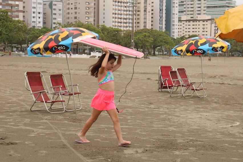 Menina caminha com prancha na faixa de areia. #pratodosverem