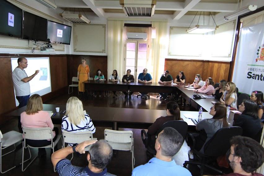 várias pessoas reunidas em torno de uma mesa em forma de U. Duas pessoas estão em pé falando ao grupo. Em uma das paredes está projetada uma imagem. #paratodosverem