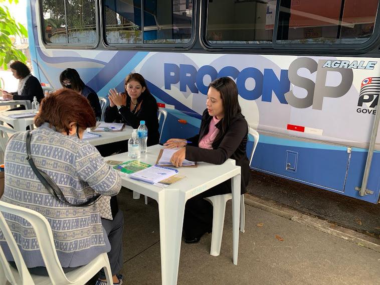 atendentes estão sentadas diante de uma ônibus onde se lê procon na lateral. Uma mulher, de costas para a foto, está sendo atendida. #paratodosverem