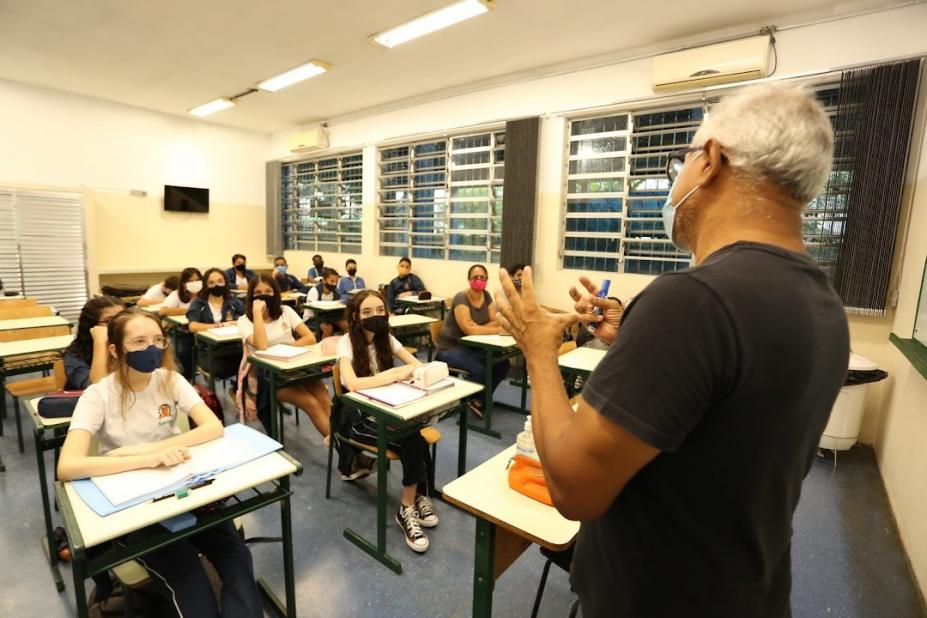 professor em primeiro plano e de costas para a foto. Alunos estão sentados assistindo a aula. #paratodosverem