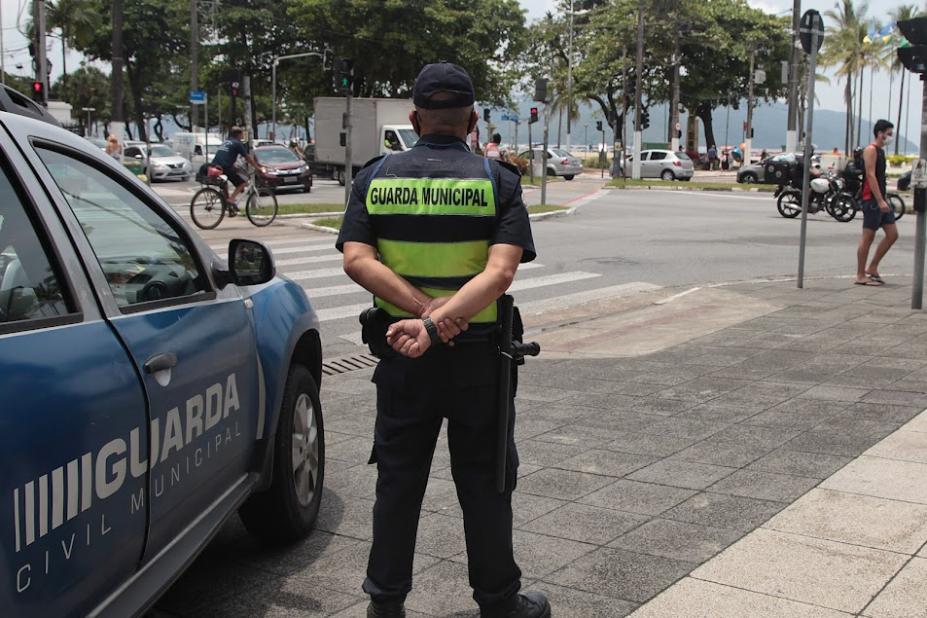 guarda municipal está de costas para foto usando colete. Ele está em pé, na orla, ao lado de uma viatura da corporação. #paratodosverem