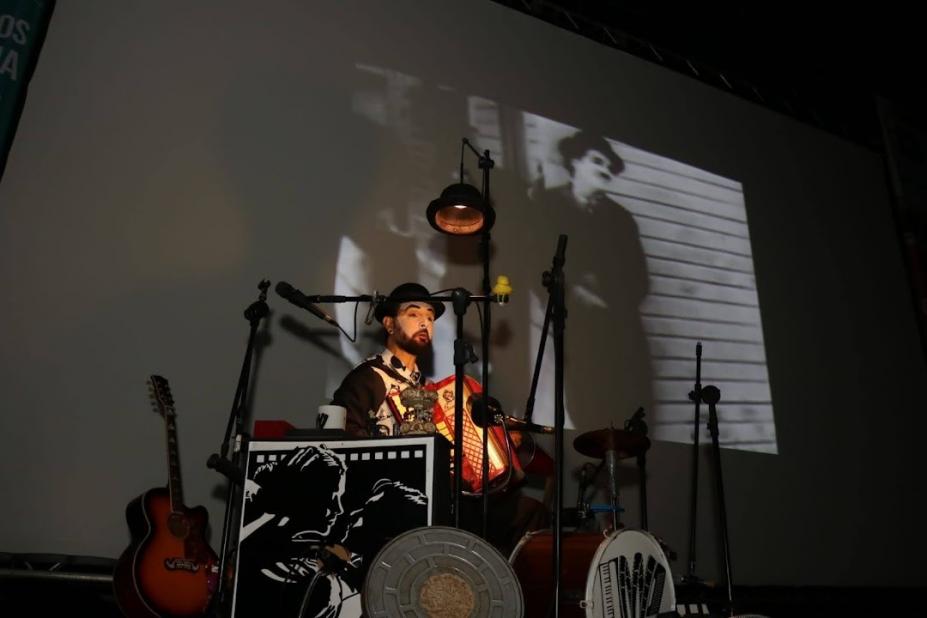 homem está tocando sanfona ao lado de outros instrumentos. Ao fundo, no telão, está projetada uma imagem de Carlitos, de Charles Chaplin. #paratodosverem