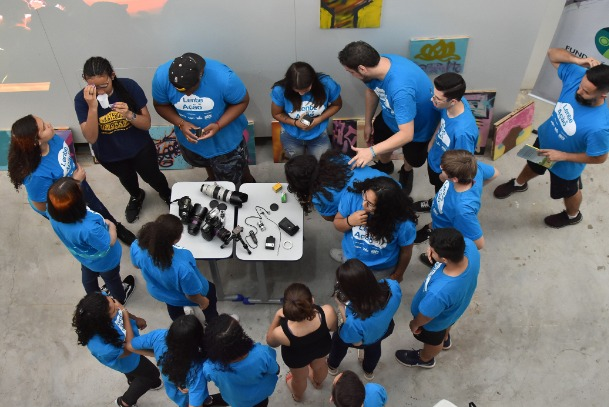 Jovens estão em pé em torno de uma mesa cheia de câmeras fotográficas. #Paratodosverem