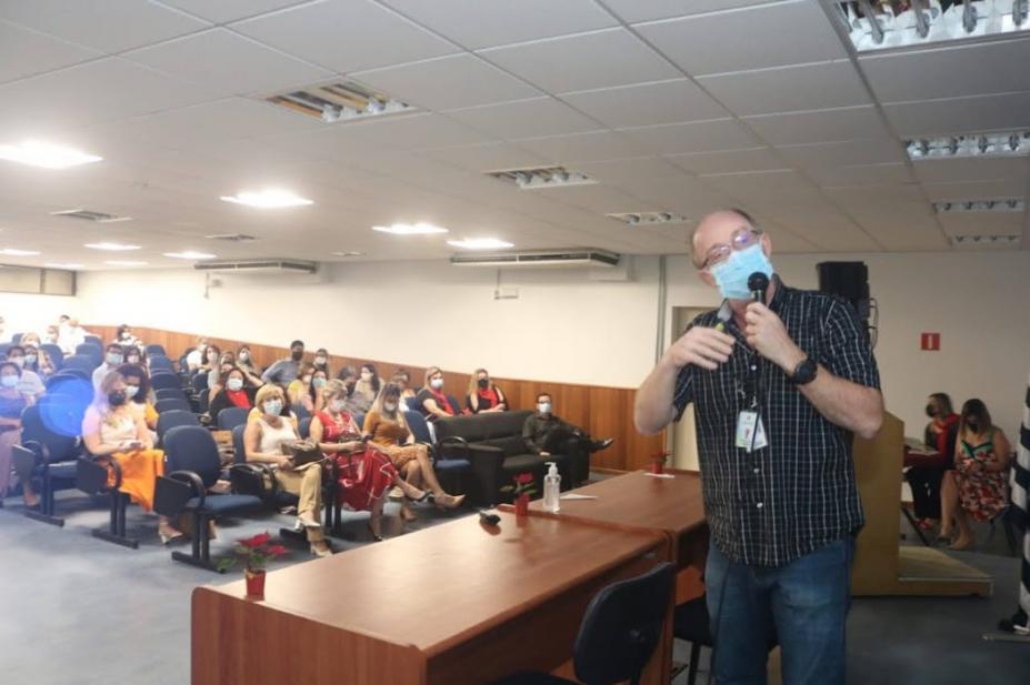 palestrante fala no microfone com público assistindo ao fundo #paratodosverem