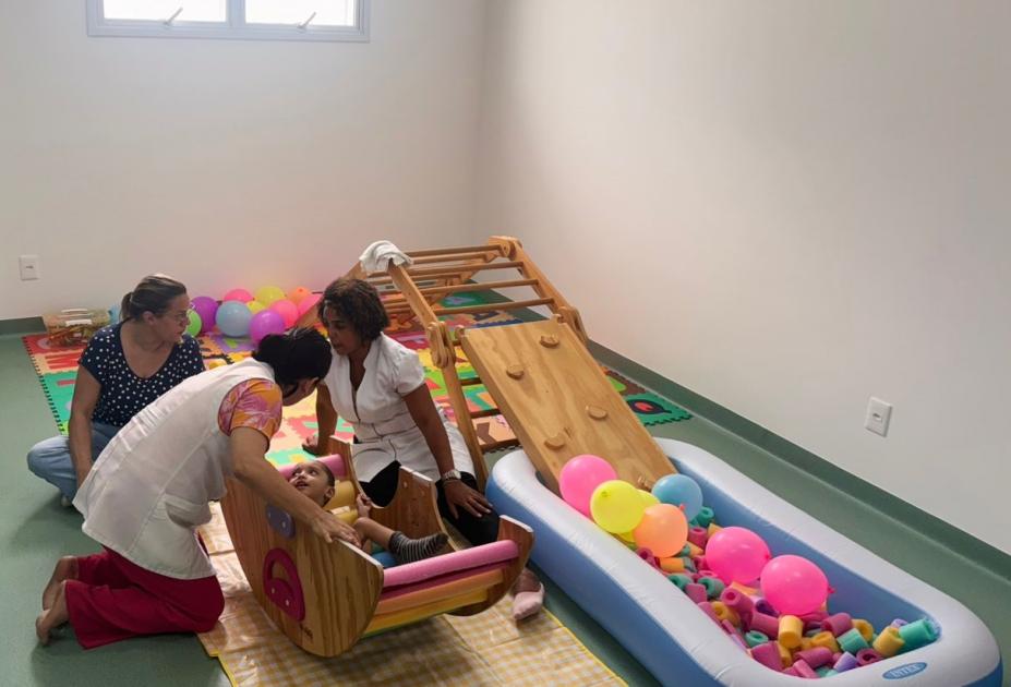 criança com comprometimento de desenvolvimento está dentro de um balanço apoiado no chão e ao lado de peças coloridas. Sob acompanhamento de três mulheres. #paratodosverem 