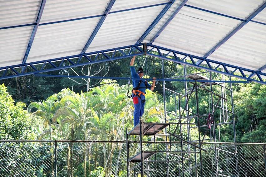 Homem está sobre andaime pintando a parte interna da estrutura da cobertura de quadra. #paratodosverem