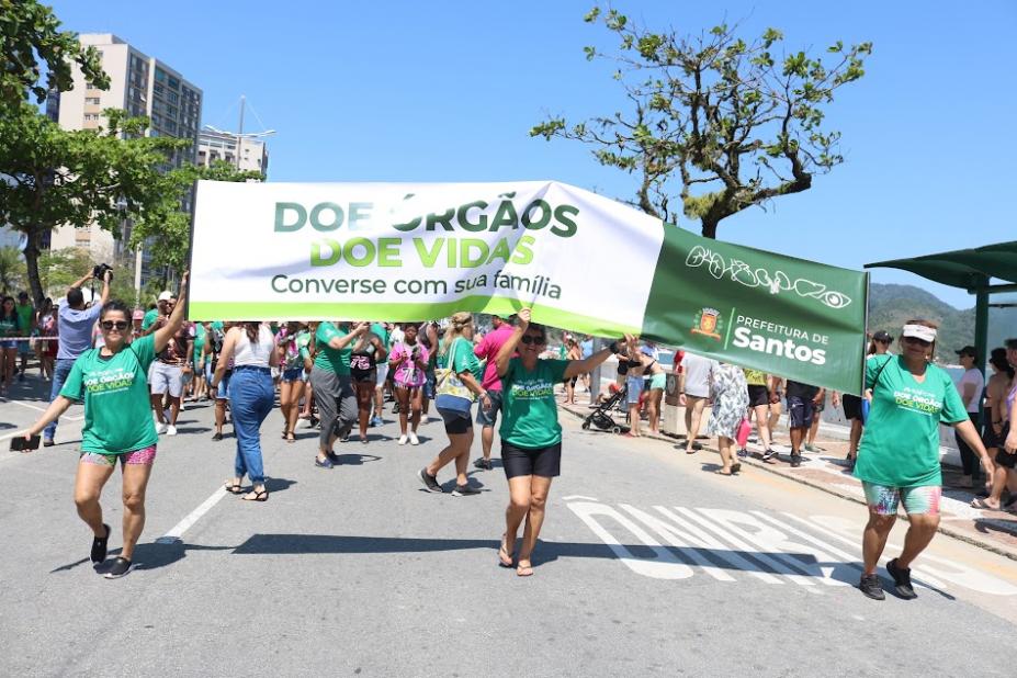 pessoas caminham em avenida segurando faixa. #paratodosverem