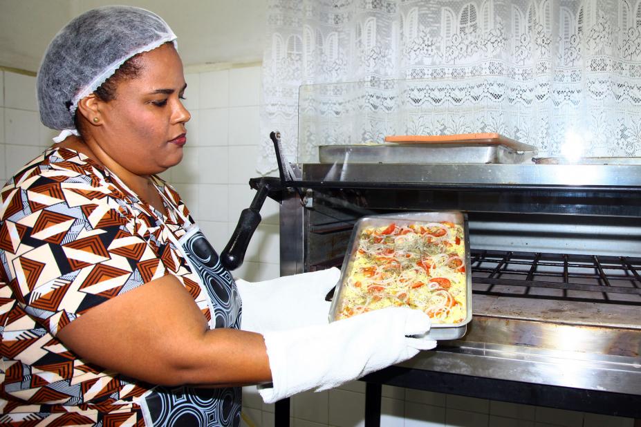 Cristiane está com luvas nas mãos. Ela retira assadeira do forno. #Pracegover