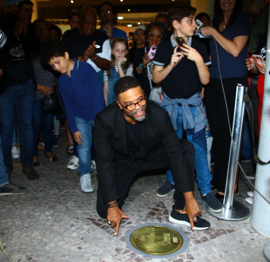 O ator santista, Luciano Quirino está agachado junto a uma estrela gravada no solo com seu nome. Há público em torno dele. #Pracegover