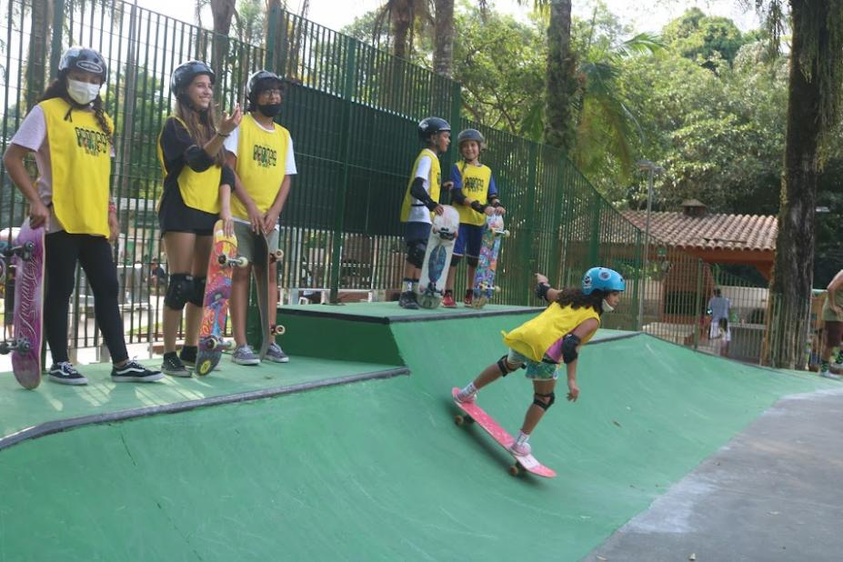 meninas estão na beira da pista aguardnado para ingressar. Uma delas já desliza com o skate na descida da rampa. A área em torno tem bastante vegetação. #paratodosverem