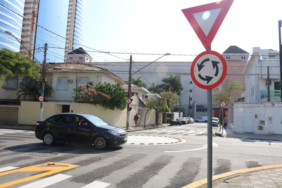 carro passa por rotatória com placa indicativa em primeiro plano #paratodosverem