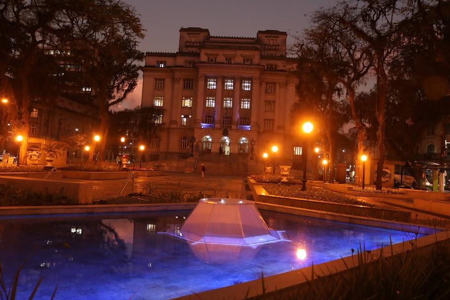 fachada da prefeitura à noite iluminada de azul.Em primeiro plano, um espelho d'água azul #paratodosverem