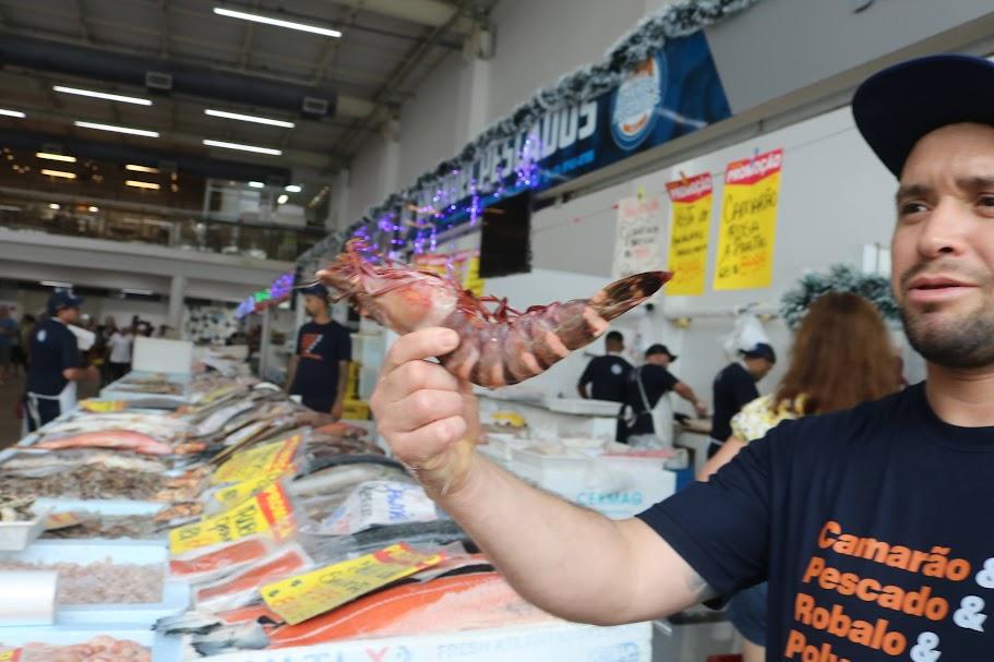 colose de camarão na mão de um vendedor. Balcões de peixe ao fundo #paratodosverem 