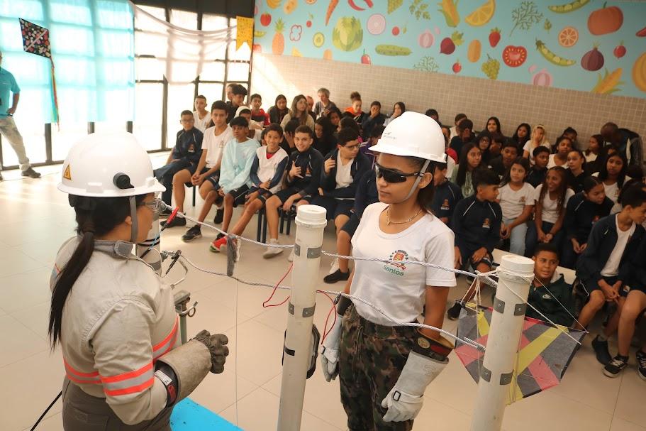 mulher uniformizada e de capacete usa maquete de fiação elétrica para demonstrar riscos com a rede elétrica. Menina à frente, com capacete e óculos, recebe orientação. #paratodosverem 