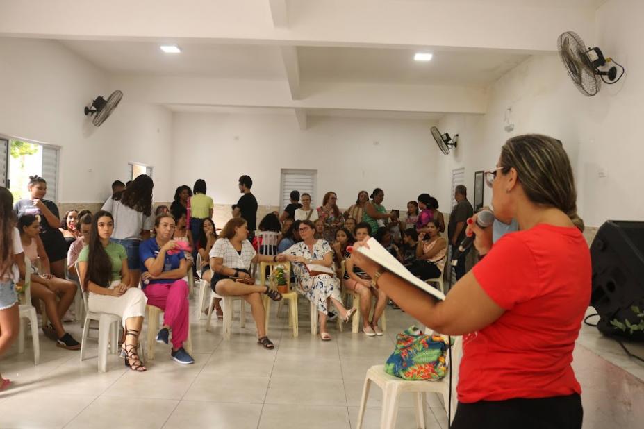 mulher está em pé com livro aberto e lendo para um grupo de outras mulheres que estão sentadas. #paratodosverem